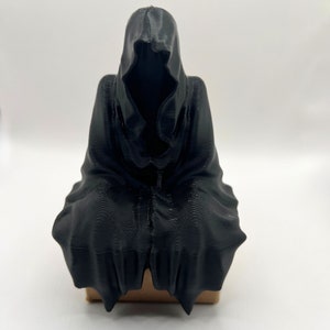 May include: A black 3D printed figurine of a hooded figure sitting with its hands clasped in its lap.