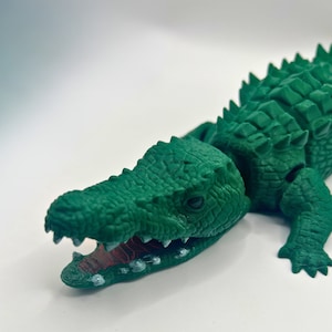 以下が含まれることがあります： 緑色のプラスチック製のワニのおもちゃ。質感のあるうろこ状の体と、鋭い歯が並んだ大きく開いた口をしています。ワニはとげのある背中で、白い表面の上に横たわっています。