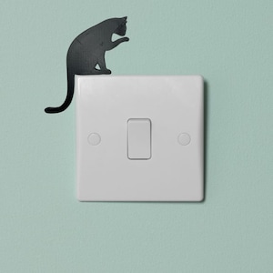 May include: A black cat silhouette sticker on a white light switch plate against a light blue wall.