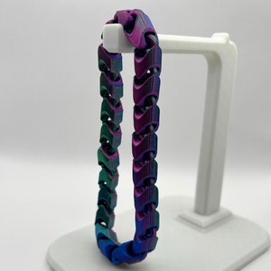 May include: A multi-coloured chain bracelet with a blue, green, and purple iridescent finish. The bracelet is made of interlocking links and is displayed on a white stand.