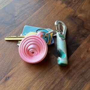 May include: A pink plastic measuring spoon keychain with a silver metal carabiner clip and a green bottle opener keychain.