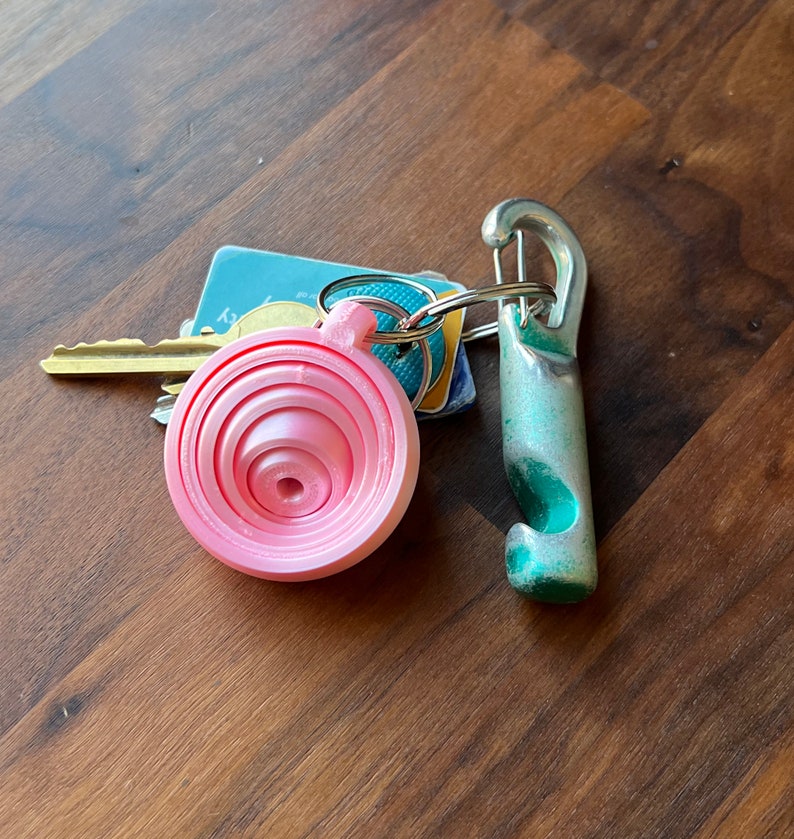May include: A pink plastic measuring spoon keychain with a silver metal carabiner clip and a green bottle opener keychain.