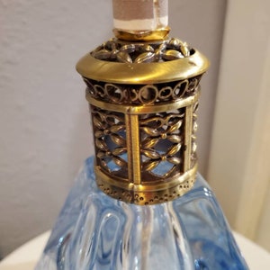 May include: A blue glass oil lamp with a gold metal top and a decorative filigree design. The lamp has a round wick and a clear glass base.