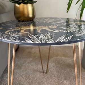 May include: A round side table with a black and gold leopard print design. The table has three gold hairpin legs.