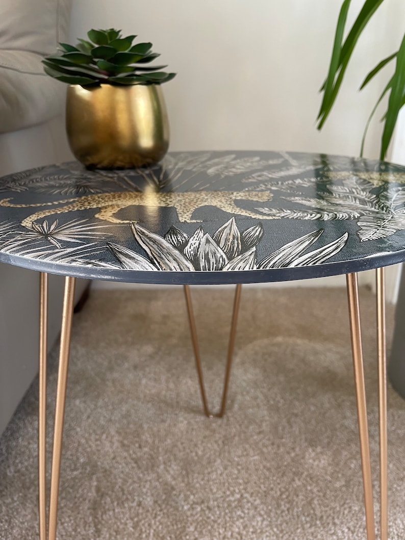 May include: A round side table with a black and gold leopard print design. The table has three gold hairpin legs.