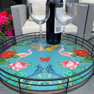 Pode incluir: Bandeja redonda com estrutura de metal preta e um design floral e de pássaros colorido sobre um fundo azul turquesa. A bandeja contém duas taças de vinho e uma garrafa de vinho tinto.