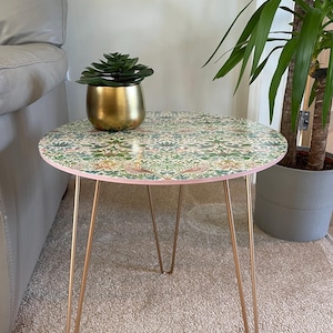 Op de afbeelding: Een ronde bijzettafel met een bloemmotief op de bovenkant en gouden haarspeldpoten. Een kleine pot met een succulent staat op de tafel.