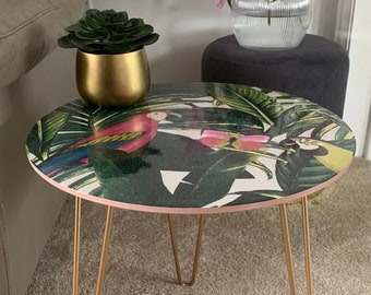 May include: A round side table with gold hairpin legs. The table top has a tropical print featuring pink and green parrots, palm leaves, and flowers.