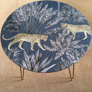 May include: A round side table with a blue fabric top featuring a white and gold tropical print with leopards. The table has gold hairpin legs.