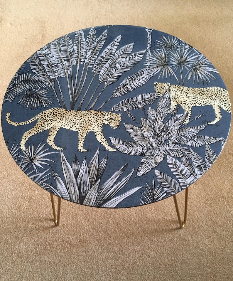 May include: A round side table with a blue fabric top featuring a white and gold tropical print with leopards. The table has gold hairpin legs.