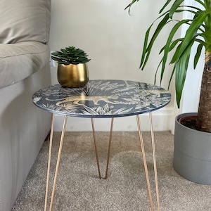 May include: A round side table with a black and gold leopard print top and gold hairpin legs. The table is sitting on a beige carpet.