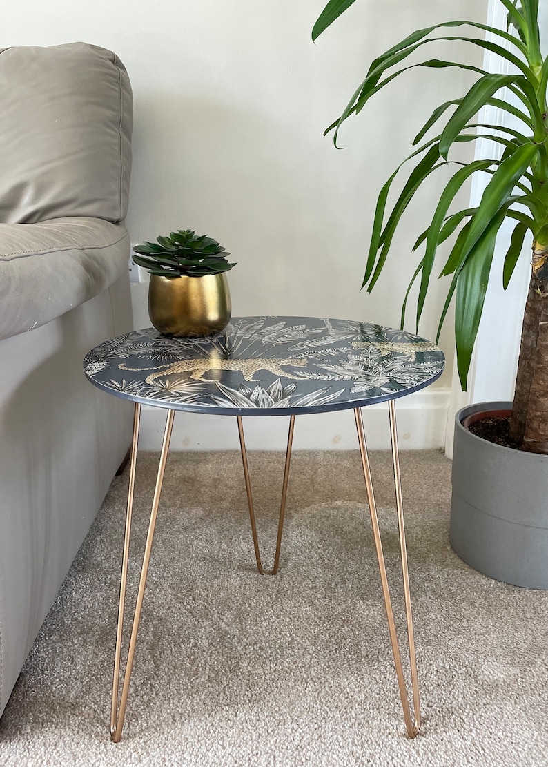 May include: A round side table with a black and gold leopard print top and gold hairpin legs. The table is sitting on a beige carpet.