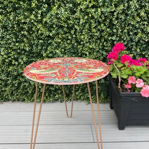 Op de afbeelding: Een ronde bijzettafel met een rood, bloemmotief tafelblad en gouden metalen haarspeldpoten. De tafel staat op een houten dek.