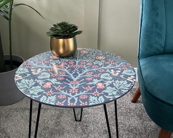 May include: A round side table with black hairpin legs. The table top has a floral pattern in shades of blue, pink, and white. A small gold pot with a green succulent plant sits on the table.