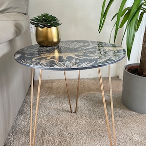 May include: A small round side table with a black and gold leopard print top and gold hairpin legs. The table is sitting on a beige carpet.