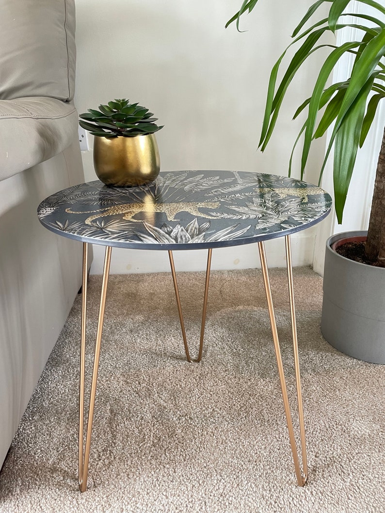 May include: A small round side table with a black and gold leopard print top and gold hairpin legs. The table is sitting on a beige carpet.