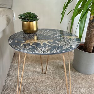 May include: A round side table with a black and white leopard print design and gold hairpin legs. The table top has a tropical leaf pattern. A small gold pot with a green succulent plant is sitting on the table.