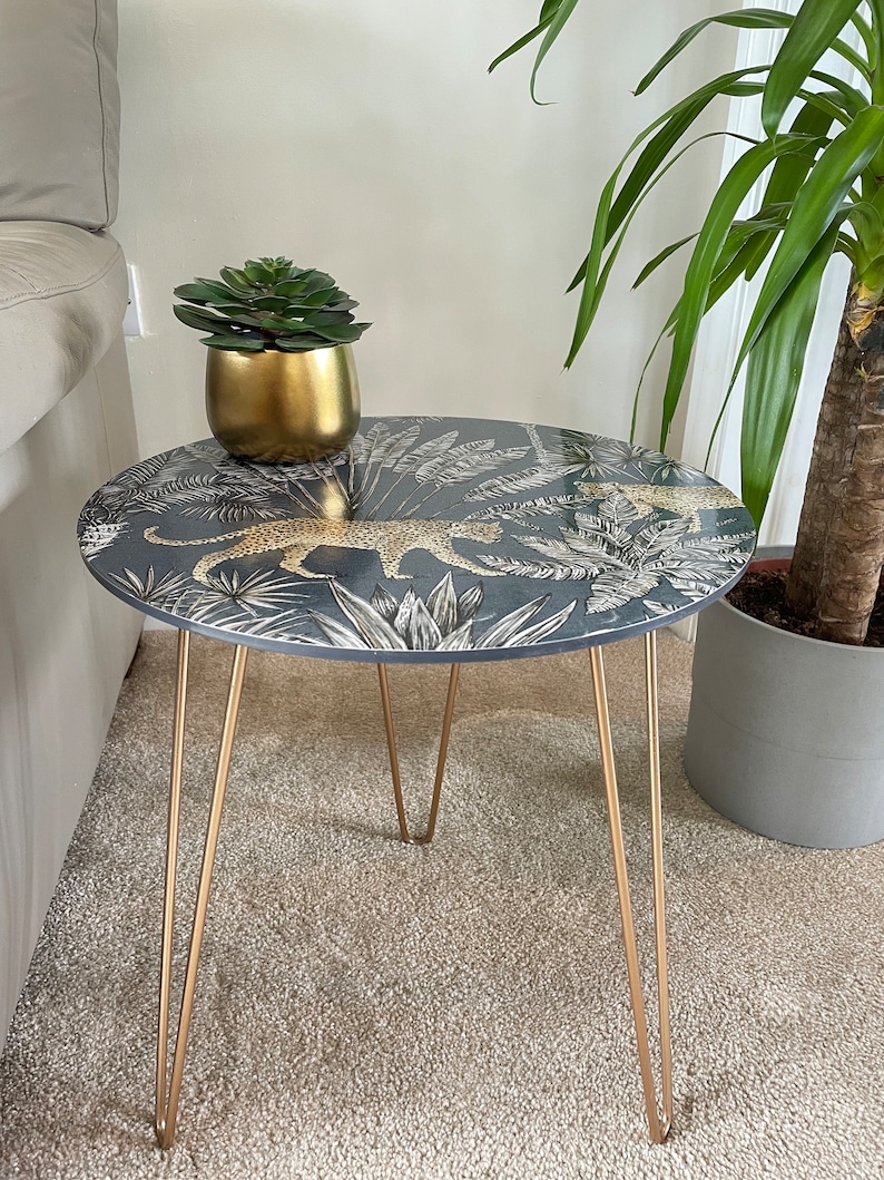 May include: A round side table with a black and white leopard print design and gold hairpin legs. The table top has a tropical leaf pattern. A small gold pot with a green succulent plant is sitting on the table.