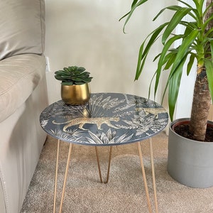 May include: A small round side table with gold hairpin legs. The table top features a dark blue background with a white and gold jungle print featuring leopards and palm leaves.