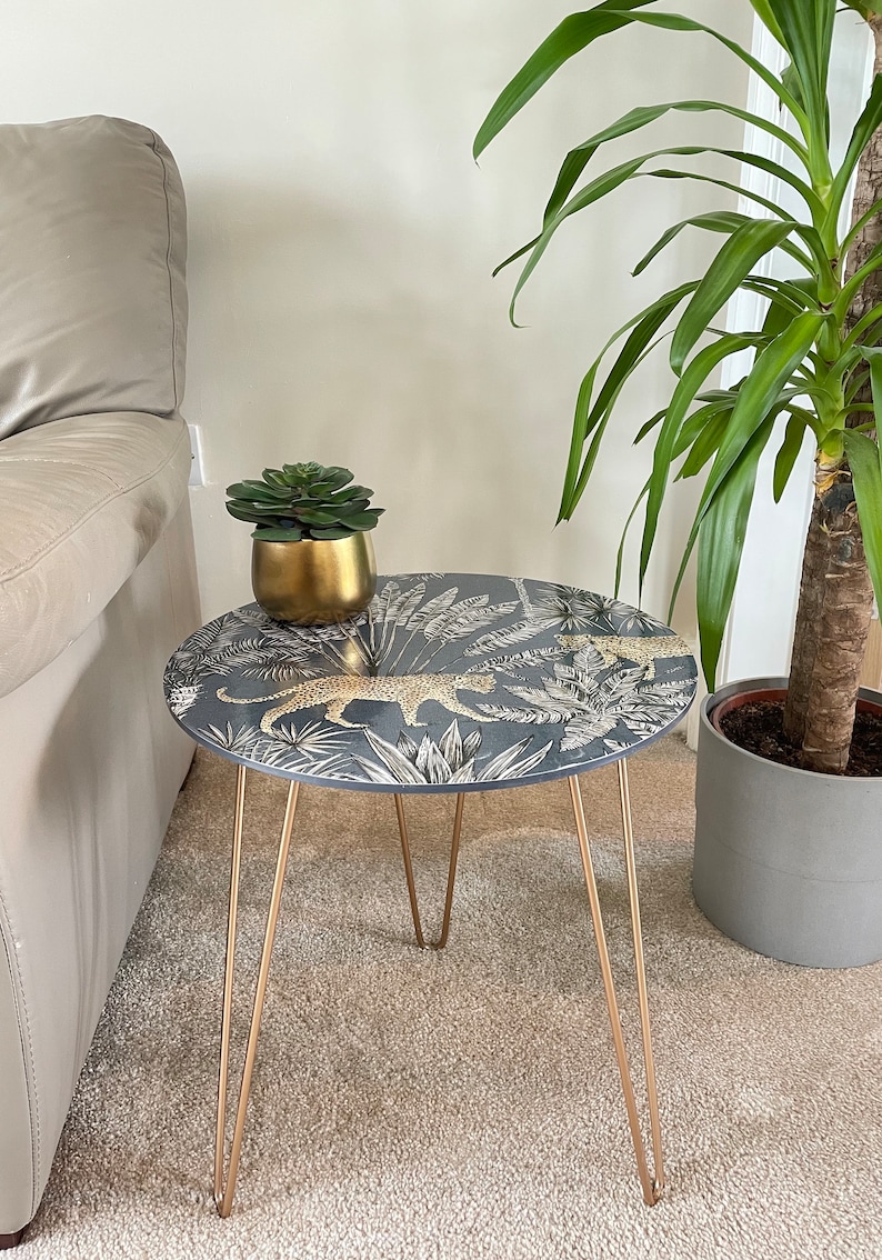 May include: A small round side table with gold hairpin legs. The table top features a dark blue background with a white and gold jungle print featuring leopards and palm leaves.