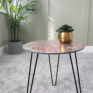 May include: A round side table with a colorful floral pattern on the top. The table has black hairpin legs and is sitting on a gray carpet.