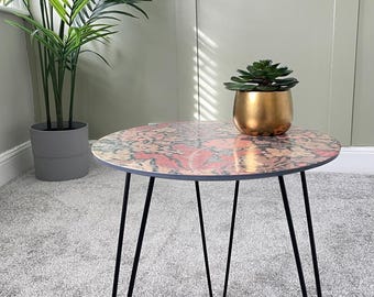 May include: A round side table with a colorful floral pattern on the top. The table has black hairpin legs and is sitting on a gray carpet.