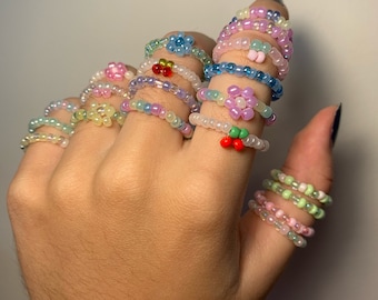 May include: A hand wearing a variety of colorful beaded rings. The rings are made with small, iridescent beads and feature different flower and fruit designs.