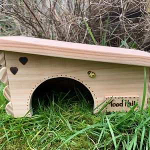 May include: A wooden toad house with a sloping roof and a small entrance hole. The house is decorated with two hearts and a small ladybug. The sign on the front reads "Toad Hall".