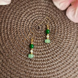 Könnte beinhalten: Ein Paar goldene Ohrringe mit grünen und weißen Perlen. Die Ohrringe liegen auf einer braunen gewebten Oberfläche.