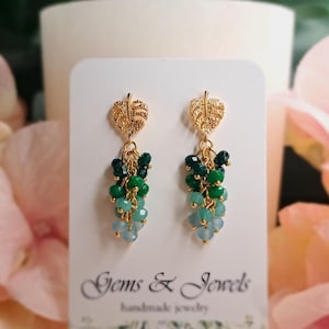Peut inclure: Boucles d'oreilles en or avec un motif de feuille et des perles vertes et bleues pendantes. Les boucles d'oreilles sont présentées sur une carte blanche avec le texte "Gems & Jewels handmade jewelry".