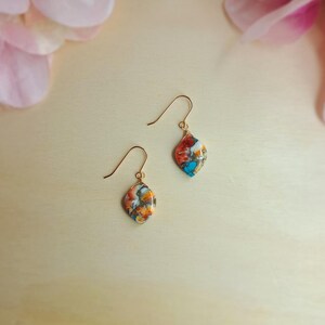 Könnte beinhalten: Ein Paar goldfarbene Ohrhänger. Jeder Ohrring hat einen bunten, oval geformten Stein mit einem Mosaikmuster aus Orange, Rot, Weiß und Türkis. Die Ohrringe werden auf einer hellen Holzoberfläche mit rosa Blumen im Hintergrund präsentiert.