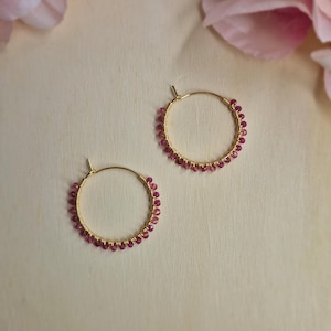 Könnte beinhalten: Ein Paar goldfarbene Creolen, verziert mit kleinen, leuchtend rosa Edelsteinen. Die Ohrringe werden auf einer hellen Holzoberfläche präsentiert, mit zarten rosa Blütenblättern im Hintergrund, was eine zarte Ästhetik erzeugt.