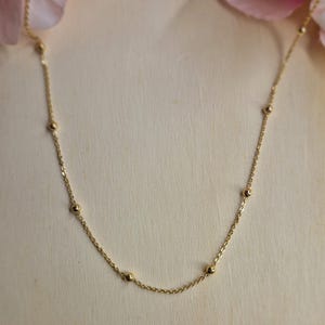Könnte beinhalten: Zarte goldene Halskette mit kleinen, gleichmäßig verteilten Perlen. Die Halskette wird vor einem weichen, neutralen Hintergrund mit rosa Blütenblättern in den oberen Ecken präsentiert. Ein schlichtes und elegantes Schmuckstück.