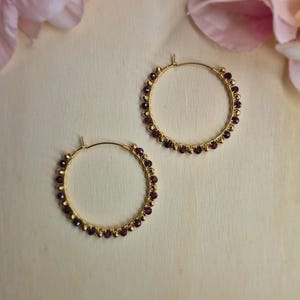 Op de afbeelding: Twee goudkleurige oorringen. Elke oorring is versierd met kleine, donkerrode kralen en goudkleurige accenten. De oorbellen worden gepresenteerd op een lichtbeige oppervlak, met roze bloemen op de achtergrond.