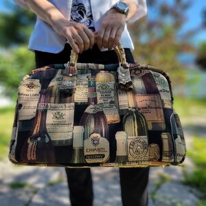 May include: A brown and beige tapestry tote bag with a wooden handle. The bag is decorated with a pattern of wine bottles and labels, including the names "Chateau Loire", "Joseph Jaca", "Chardonnay", "Chateau Blanc", "Chianti", and "Tuscany".