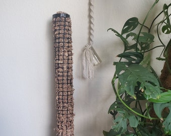 May include: A brown moss pole with a black wire mesh covering. The pole is standing upright on a black surface. A potted plant with large green leaves is in the background.