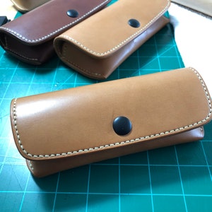 以下が含まれることがあります： 白いステッチと黒いボタンが付いた茶色の革製のケースが3つ。ケースは緑色のカッティングマットの上に置かれています。