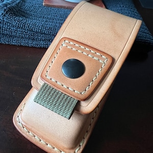 Puede incluir: Una funda de cuero marrón claro con cierre de presión y una correa verde. La funda tiene costuras blancas alrededor de los bordes y un botón negro en la parte delantera.