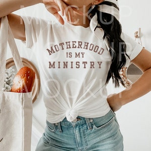May include: A white t-shirt with a brown graphic that says "Motherhood is my Ministry". The shirt is tied at the front and the model is wearing blue jeans.