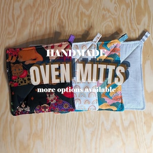 May include: A selection of handmade oven mitts with diverse patterns, such as cats, rainbows, and geometric shapes. The text "HANDMADE OVEN MITTS" and "more options available" are visible. Each mitt includes a hanging loop.