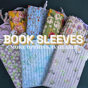 May include: A collection of quilted fabric book sleeves featuring various floral and botanical patterns in yellow, blue, green, purple, and beige. Each rectangular sleeve includes a fabric tie closure and the text BOOK SLEEVES MORE OPTIONS AVAILABLE.