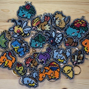 Puede incluir: Una colección de llaveros de personajes pixelados en varios colores, incluyendo azul, naranja, amarillo y verde. Cada llavero presenta un diseño de personaje diferente con un llavero plateado adjunto. Los llaveros están dispuestos sobre una superficie de madera marrón claro.