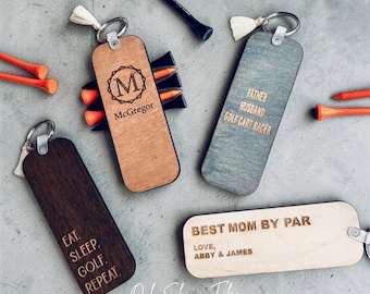 May include: Wooden keychains with golf-themed phrases and names. One keychain says "Eat. Sleep. Golf. Repeat." Another says "Best Mom by Par, Love, Abby & James." Other keychains have the text "McGregor" and "Father, Husband, Golf Cart Racer."