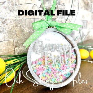 Puede incluir: Un adorno de Pascua en forma de conejo de purpurina plateada con una ventana de plástico transparente llena de chispas de colores. El adorno dice "Bunny Bait" en letras de purpurina plateada.