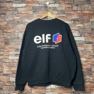 Op de afbeelding: Zwarte longsleeve sweater met de tekst "elf" in witte letters en een rood, wit en blauw logo. De tekst "Excitement Staff International" is onder het logo gedrukt.