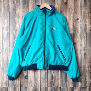 Könnte beinhalten: Eine tealblaue Fleecejacke mit Reißverschluss und einem schwarzen und tealblauen Kragen. Die Jacke hat zwei Reißverschlusstaschen auf der Vorderseite und ein kleines Logo-Patch auf der linken Brust.