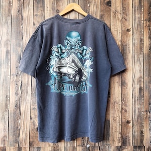 May include: A dark gray t-shirt featuring a graphic of a clown with a car and the text "The Puppet Master" in a blue neon style. The shirt is hanging on a wooden hanger against a wooden background.