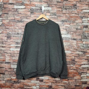 Könnte beinhalten: Ein dunkelgraues Sweatshirt hängt an einem Holzbügel vor einer Backsteinmauer. Das Sweatshirt mit Rundhalsausschnitt hat lange Ärmel und ein kleines Detail am linken Ärmel. Der Stoff scheint ein weiches, bequemes Material zu sein.