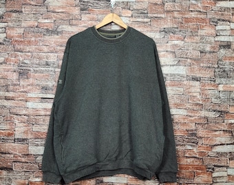 May include: A dark gray sweatshirt hanging on a wooden hanger against a brick wall. The sweatshirt has a crew neck and long sleeves. There is a small detail on the left sleeve. The fabric appears to be a soft, comfortable material.