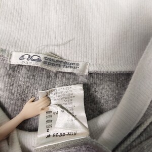 May include: A gray and white long-sleeve shirt with a white collar and a white tag that reads "Courr&egrave;ges Sport futur". The tag is attached to the shirt with a white thread. The shirt has a white label with the text "ATN-0096 W" and a size tag with the text "2".
